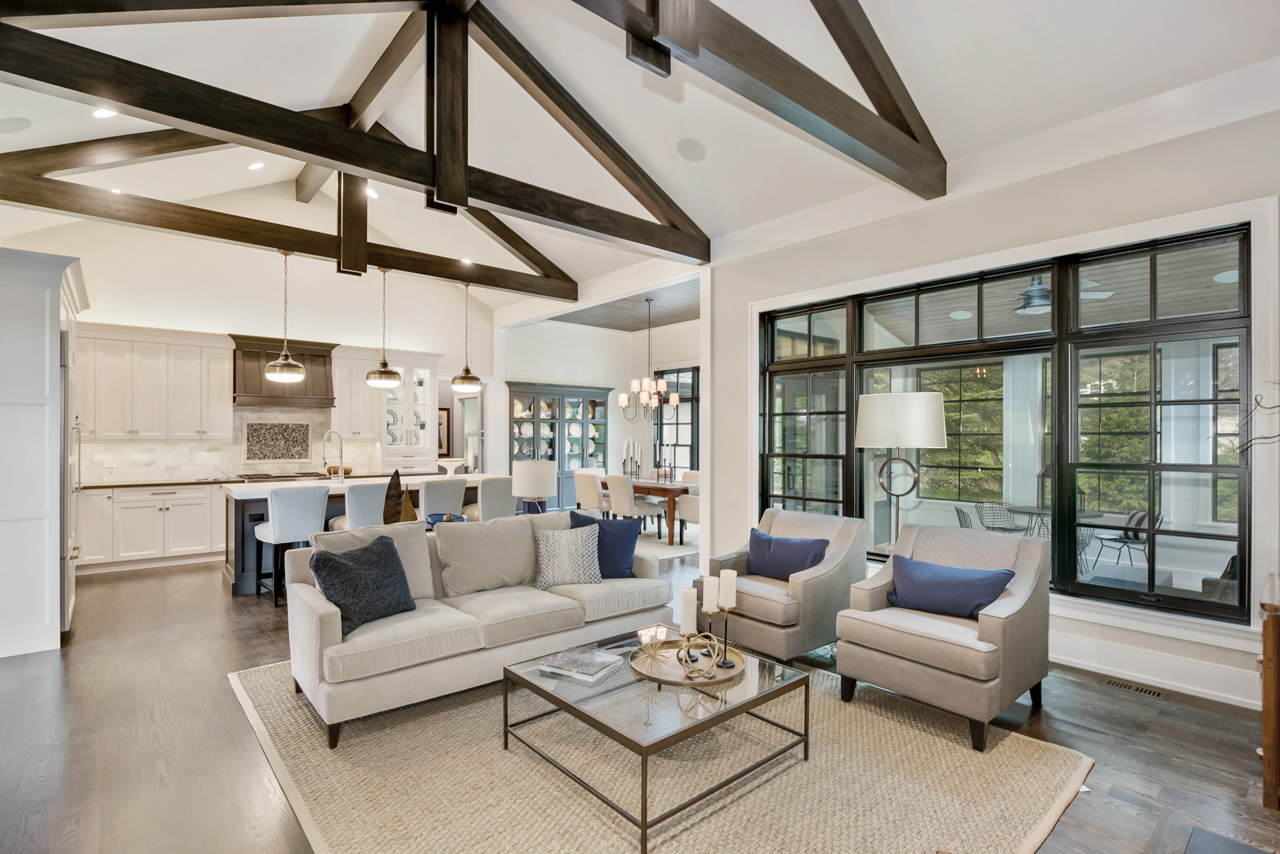 A bright, open-concept living area with light wood flooring, a minimalist staircase with wooden steps and white risers, and a cozy seating area with a gray sofa. Features include a built-in fireplace, a flat-screen TV, a potted plant, and large windows that let in natural light.