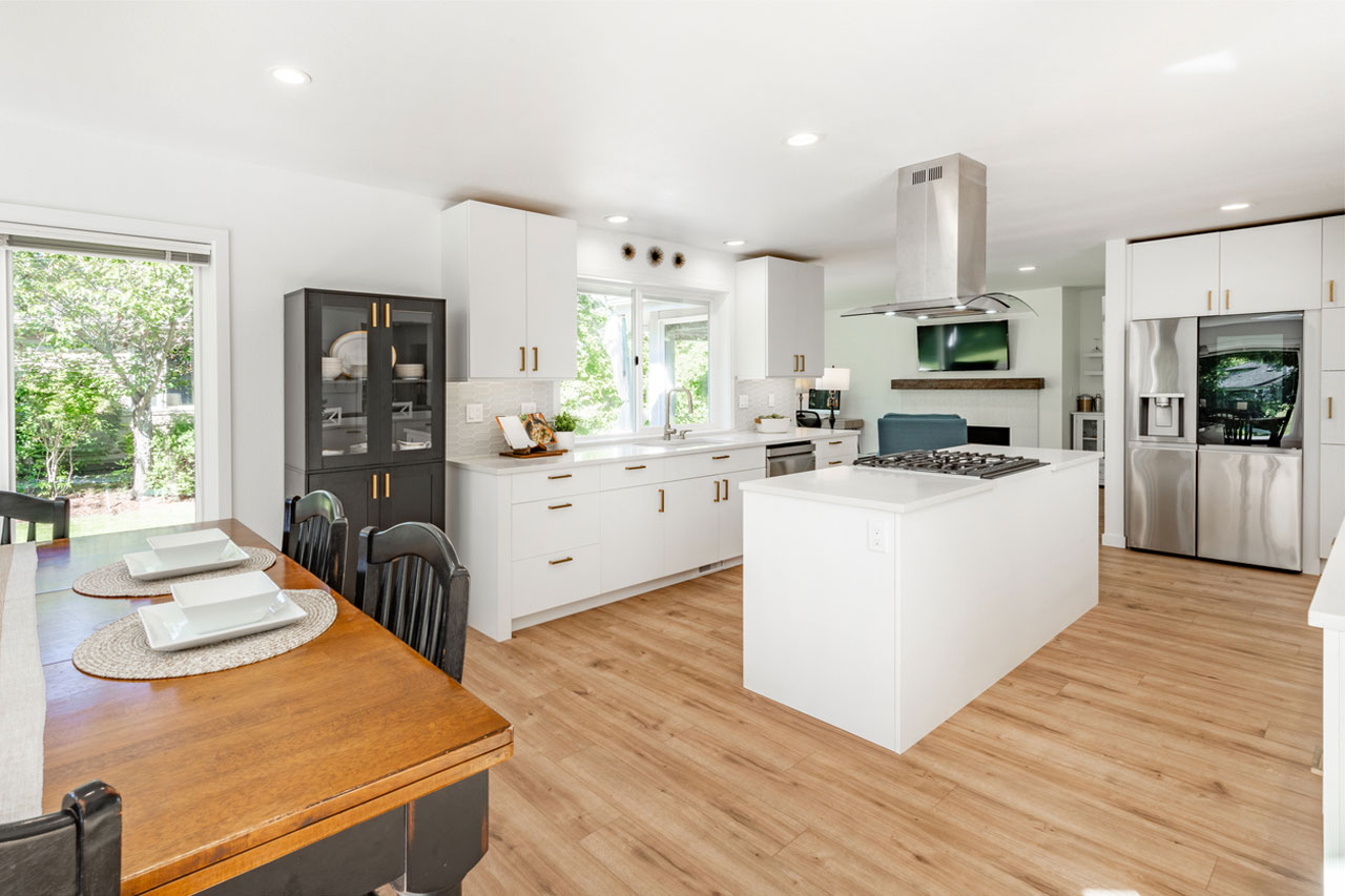 A contemporary kitchen with white cabinets, gold hardware, a large white center island, and stainless steel appliances including a built-in refrigerator and range hood. The room features light wood flooring, a wooden dining table with black chairs, and a window offering a view of greenery outside.